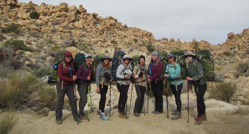 a group of veterans carrying large backpacks and trekking poles pose for a photo in front of a rocky landscape in joshua tree